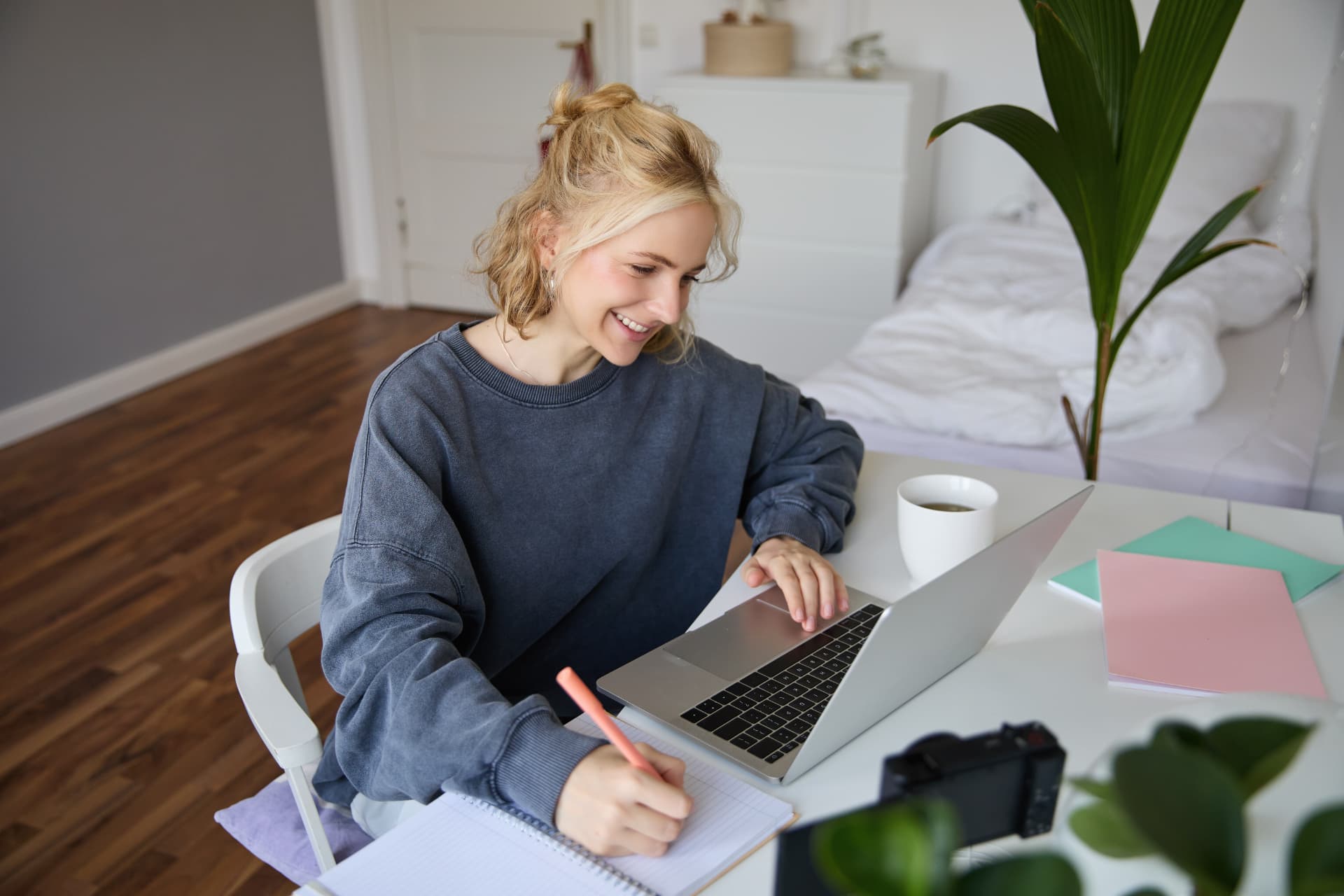 A smiling young woman studies at her desk, writing in a notebook beside an open laptop